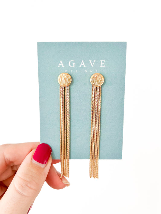 A hand adorned with red nail polish holds a card from Agave Designs, displaying the elegant Tassel Studs. These 14k gold plated earrings feature textured circular tops and multiple fine chains measuring 80mm x 10mm. The plain white background accentuates their luxurious appeal.