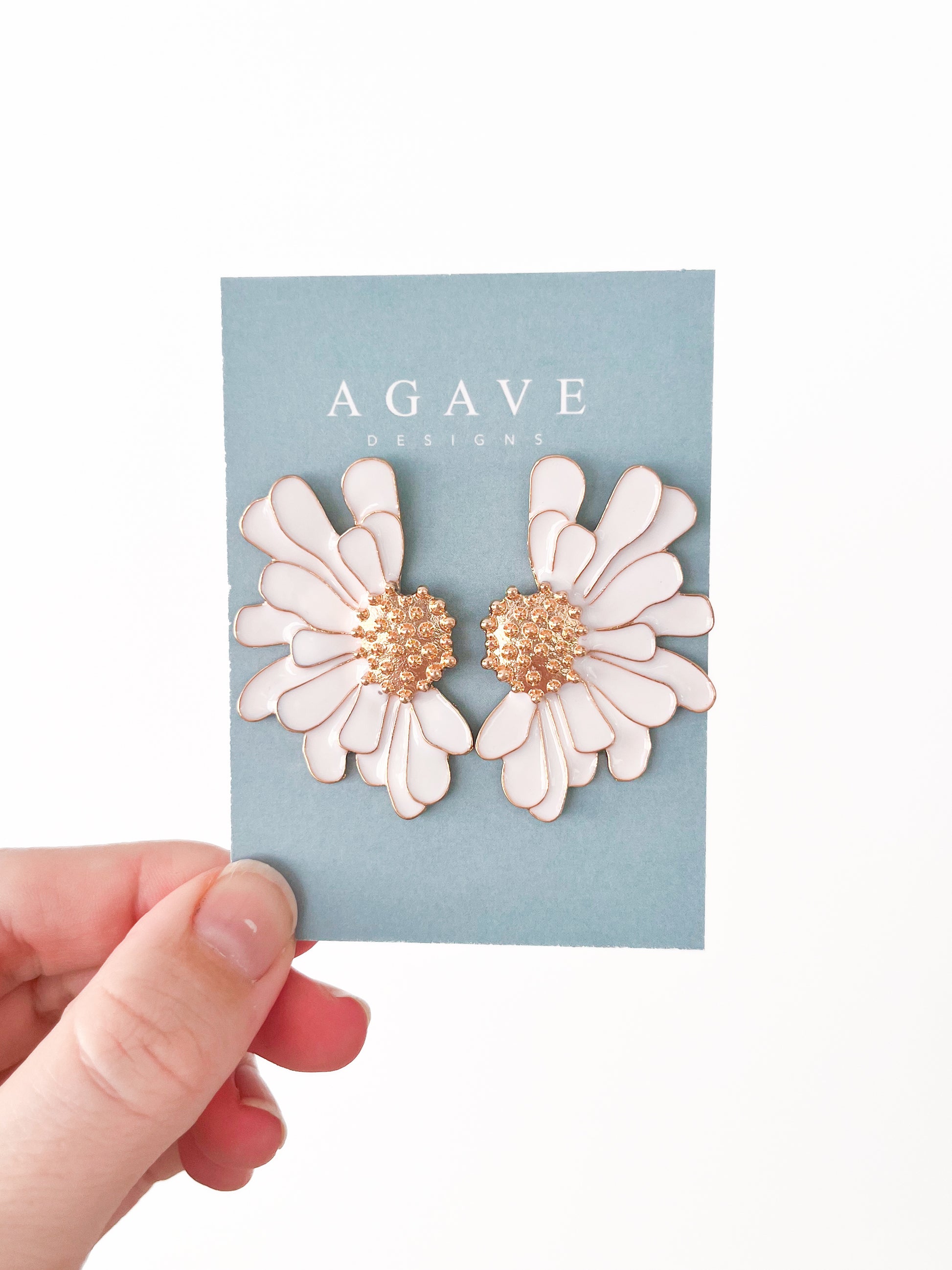 A hand holds a sleek card displaying "Agave Designs," showcasing two exquisite White ‘n Gold Flower Studs with radiant gold centers. The jewelry shines vividly against the plain white background.