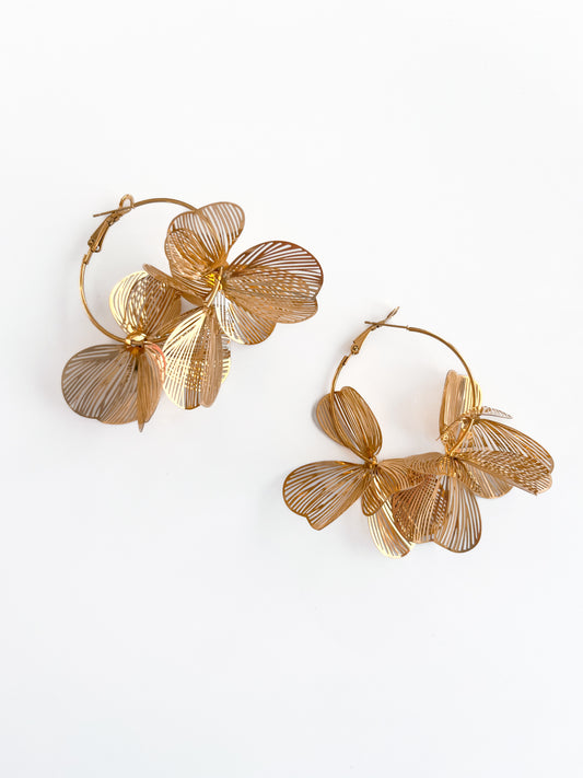 The Gold Flower Hoops by Agave Designs are intricately designed gold wire hoop earrings that feature delicate leaf-like patterns resembling flowers. These are crafted in durable stainless steel and set against a plain white background.