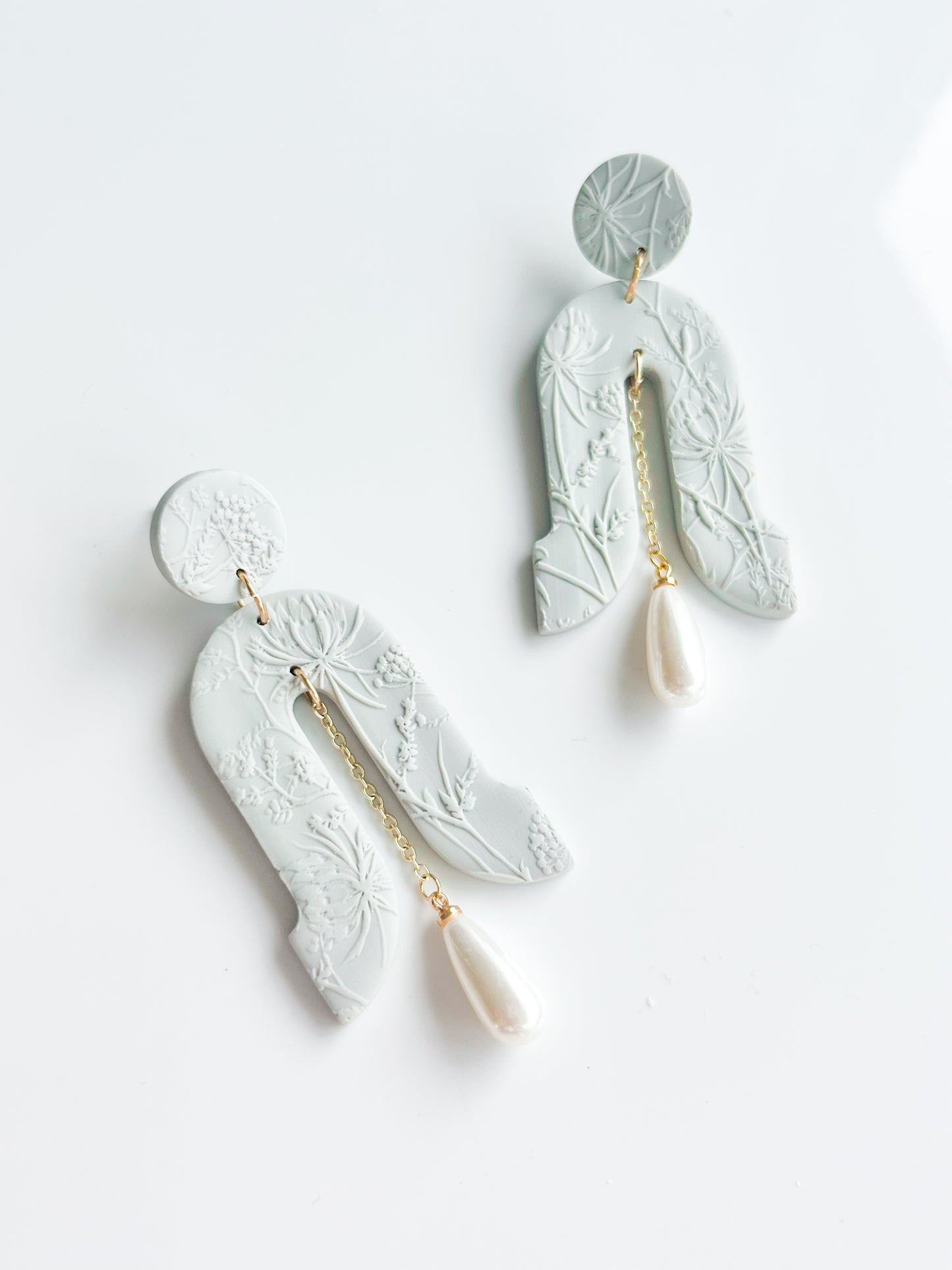 The Gloria earrings by Agave Designs feature light gray textured polymer clay with floral patterns, gold chains, and white teardrop pearls on sterling silver hooks, shown on a white background.