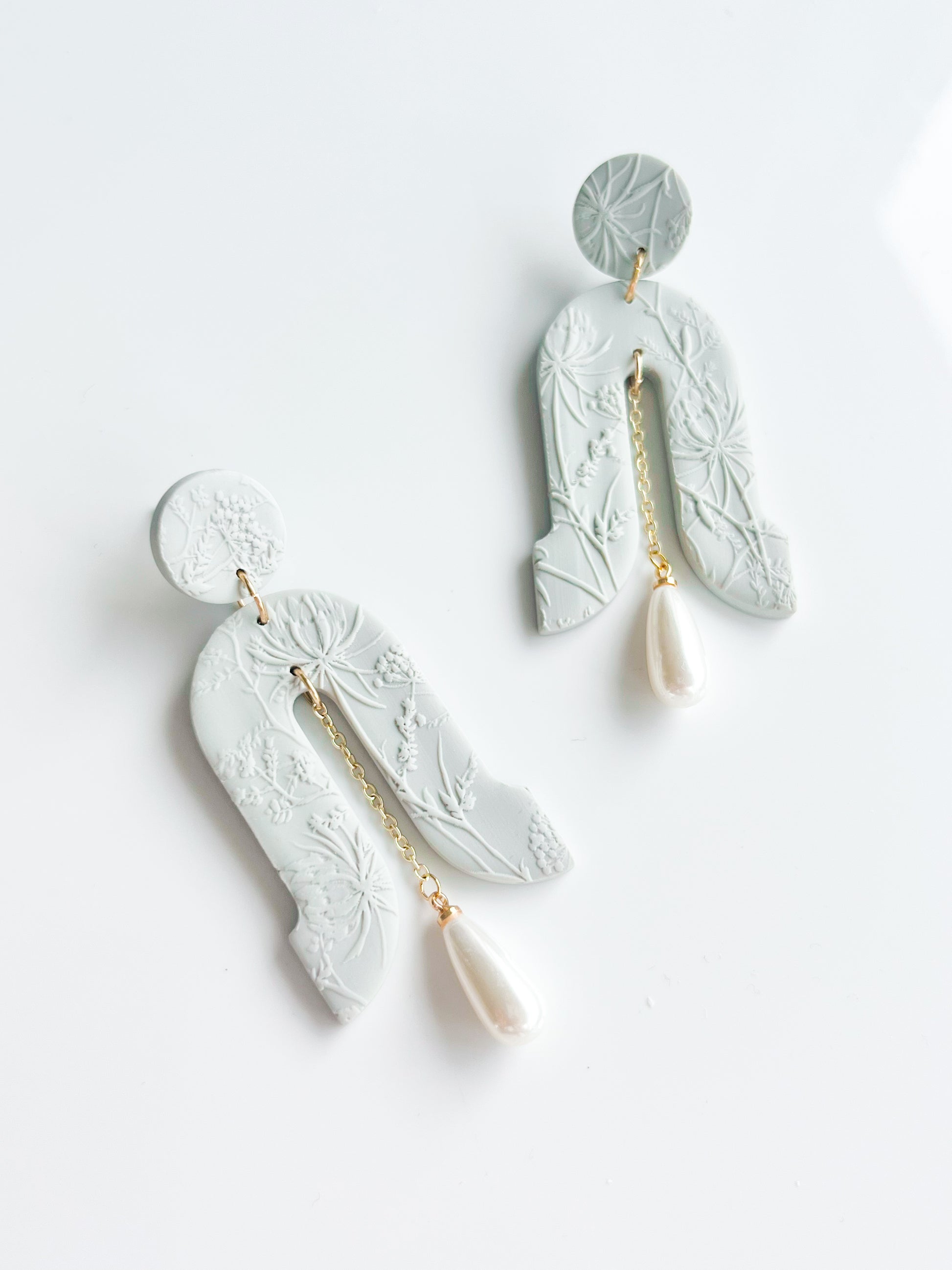 The Gloria earrings by Agave Designs feature light gray textured polymer clay with floral patterns, gold chains, and white teardrop pearls on sterling silver hooks, shown on a white background.