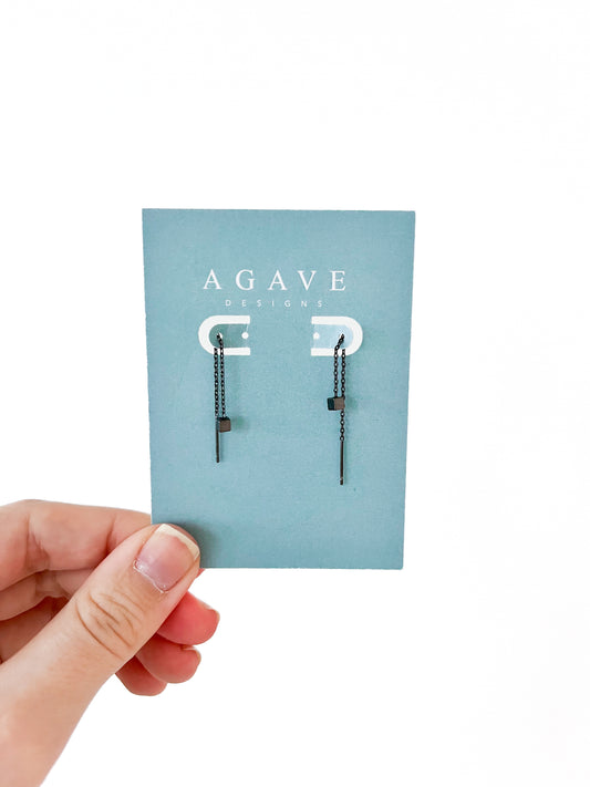 A hand holds a card labeled "Agave Designs," showcasing the Little Square Danglies Black earrings. These minimalist geometric earrings are crafted from zinc alloy, featuring a gold linear design with delicate chains and rectangular shapes, set against a blue-green background.