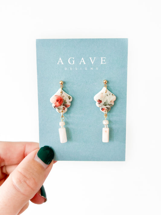 A hand with green painted nails holds up a light blue card labeled "Agave Designs," displaying a pair of the Fall Romantic Bead Danglies. The earrings, featuring floral-patterned ceramic pieces and rectangular white stones, are elegantly gold plated. The background is white.