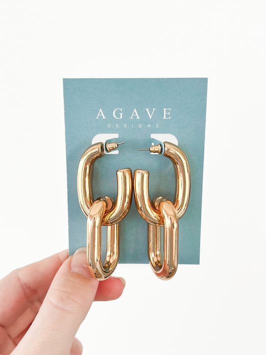 A hand displays a card featuring the Link Hoops, a pair of chunky gold chain-link earrings made from zinc alloy by Agave Designs, set against a plain white background.