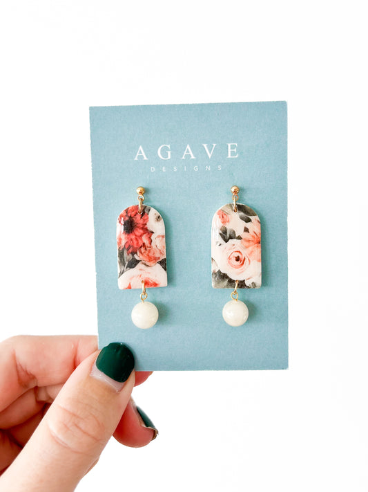 A hand with dark green nail polish holds a light blue card displaying the Fall bead Danglies from Agave Designs. These floral earrings showcase a pattern with whites, pinks, and reds, and are complemented by a round white bead at the bottom.