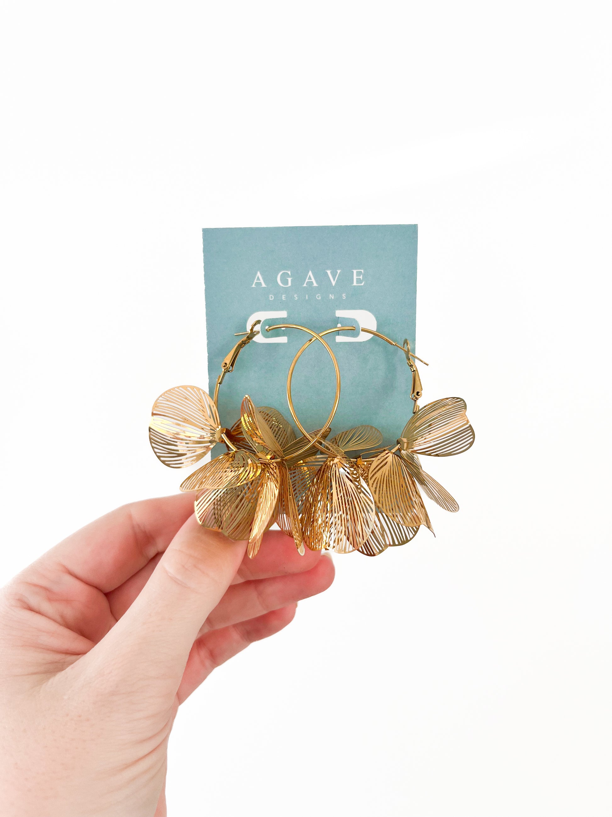 A hand holds the Gold Flower Hoops by Agave Designs, crafted in stainless steel and featuring leaf-shaped charms, displayed against a light blue card labeled "AGAVE DESIGNS.