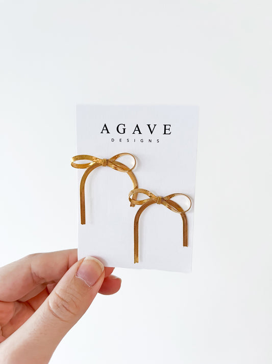 A hand presents a "Little Bow Studs" card from Agave Designs, showcasing elegant gold, stainless steel bow-shaped earrings against a plain white background.