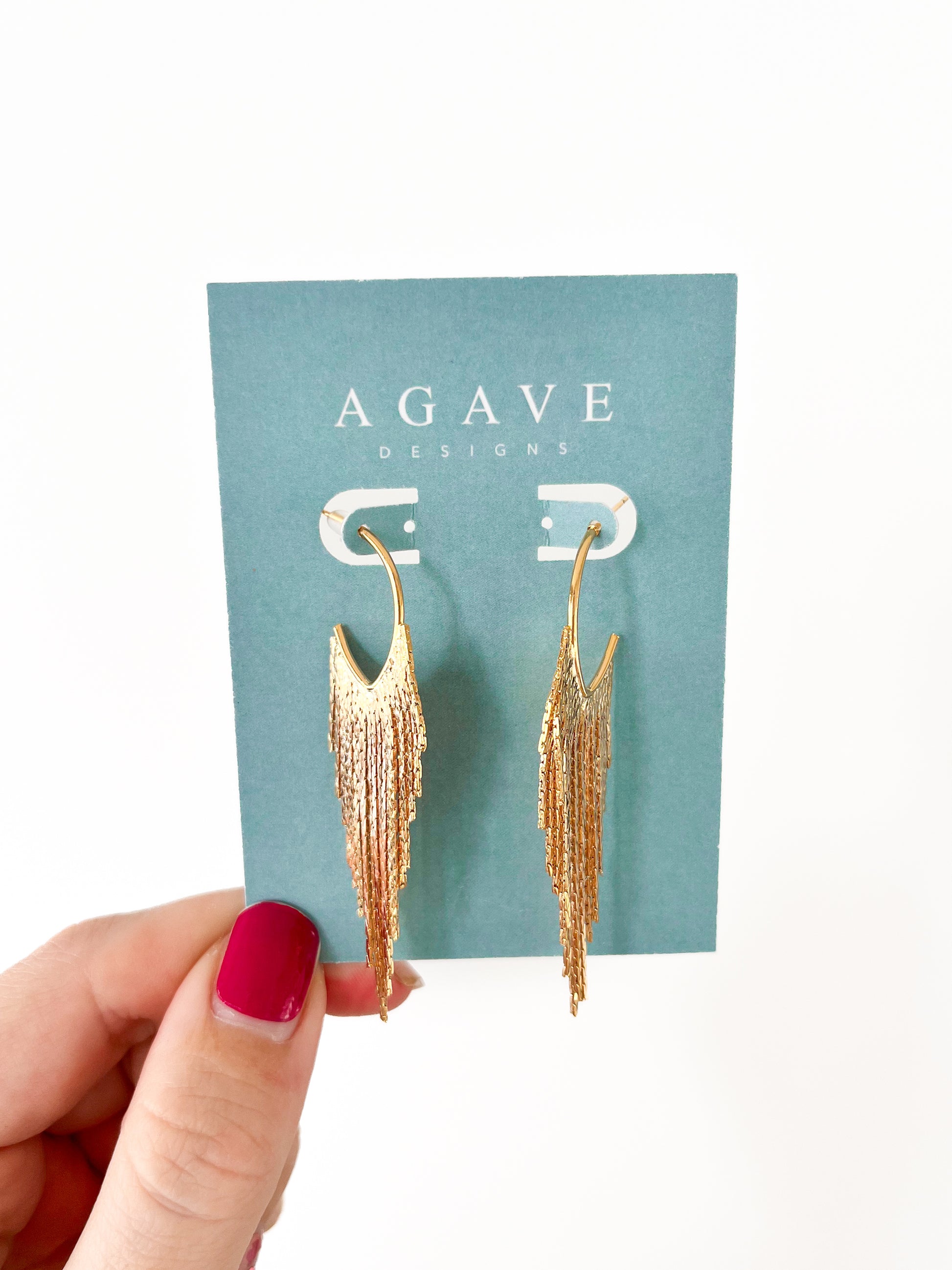 A hand with red nail polish holds a card displaying "Gold Feathers," 14k gold plated earrings by Agave Designs, against a turquoise background. The textured, cascading earrings measure 65mm x 18mm.