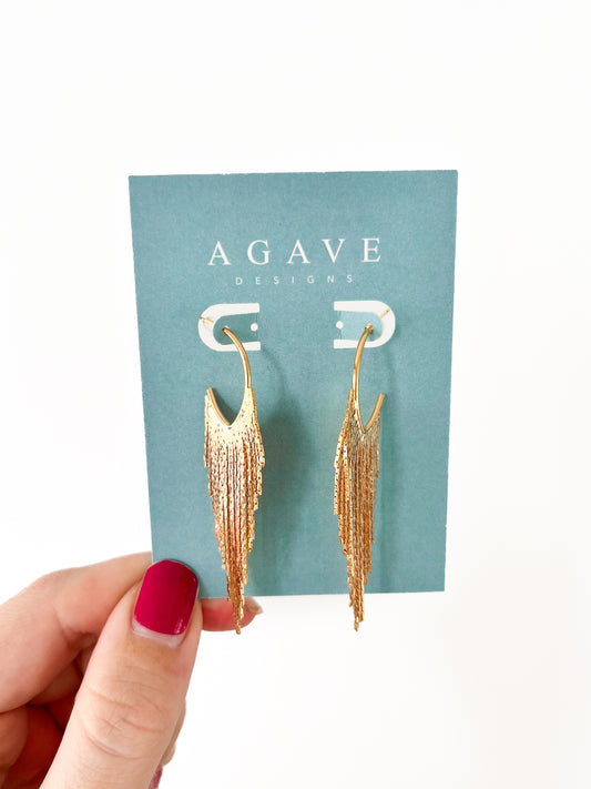 A hand with red nail polish holds a card displaying "Gold Feathers," 14k gold plated earrings by Agave Designs, against a turquoise background. The textured, cascading earrings measure 65mm x 18mm.
