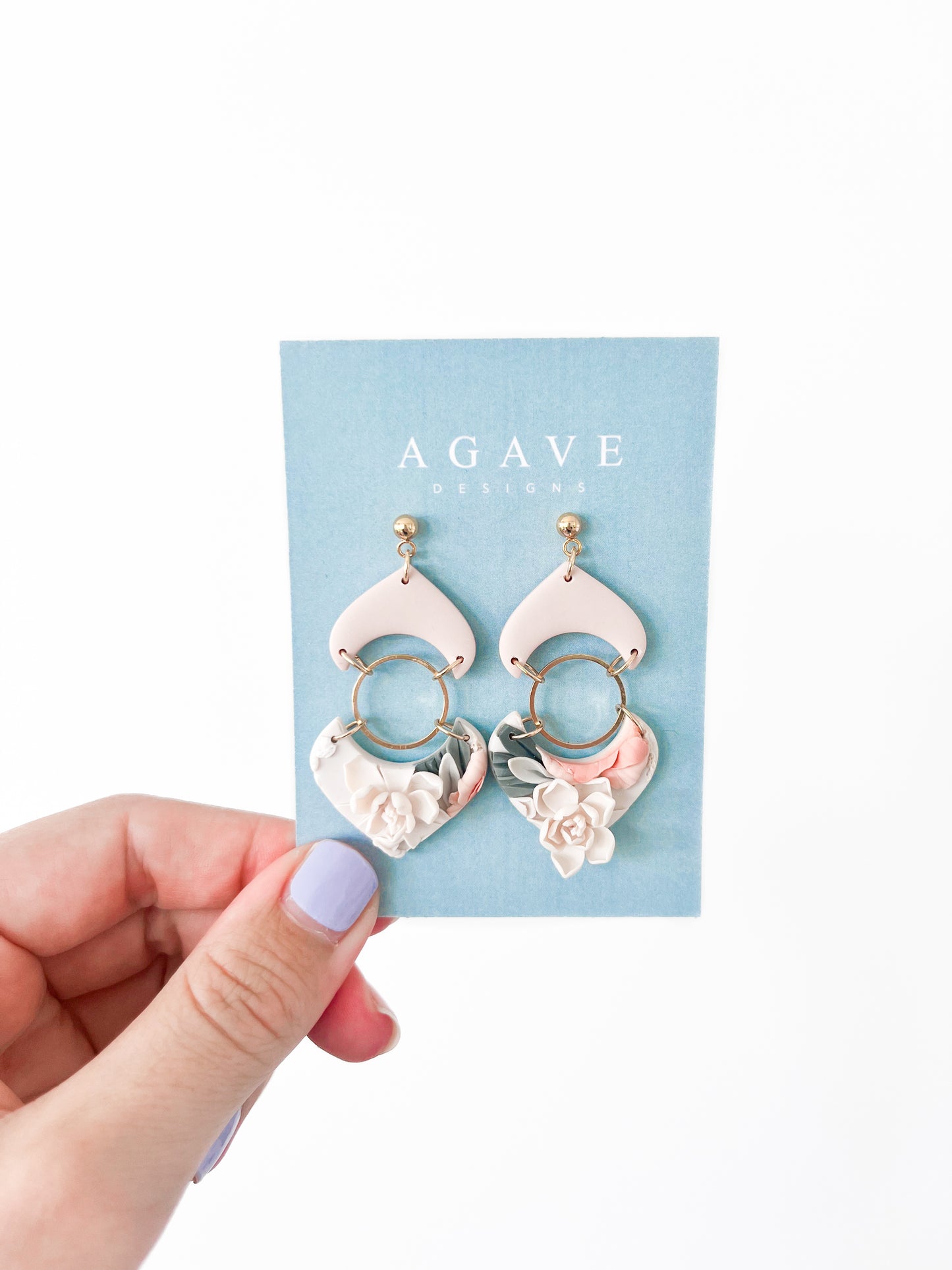 A hand with lavender-painted nails holds a blue "Agave Designs" card displaying the Sarah earrings—gold-plated, made to order statement jewelry with pink and gold dangles and white floral accents.
