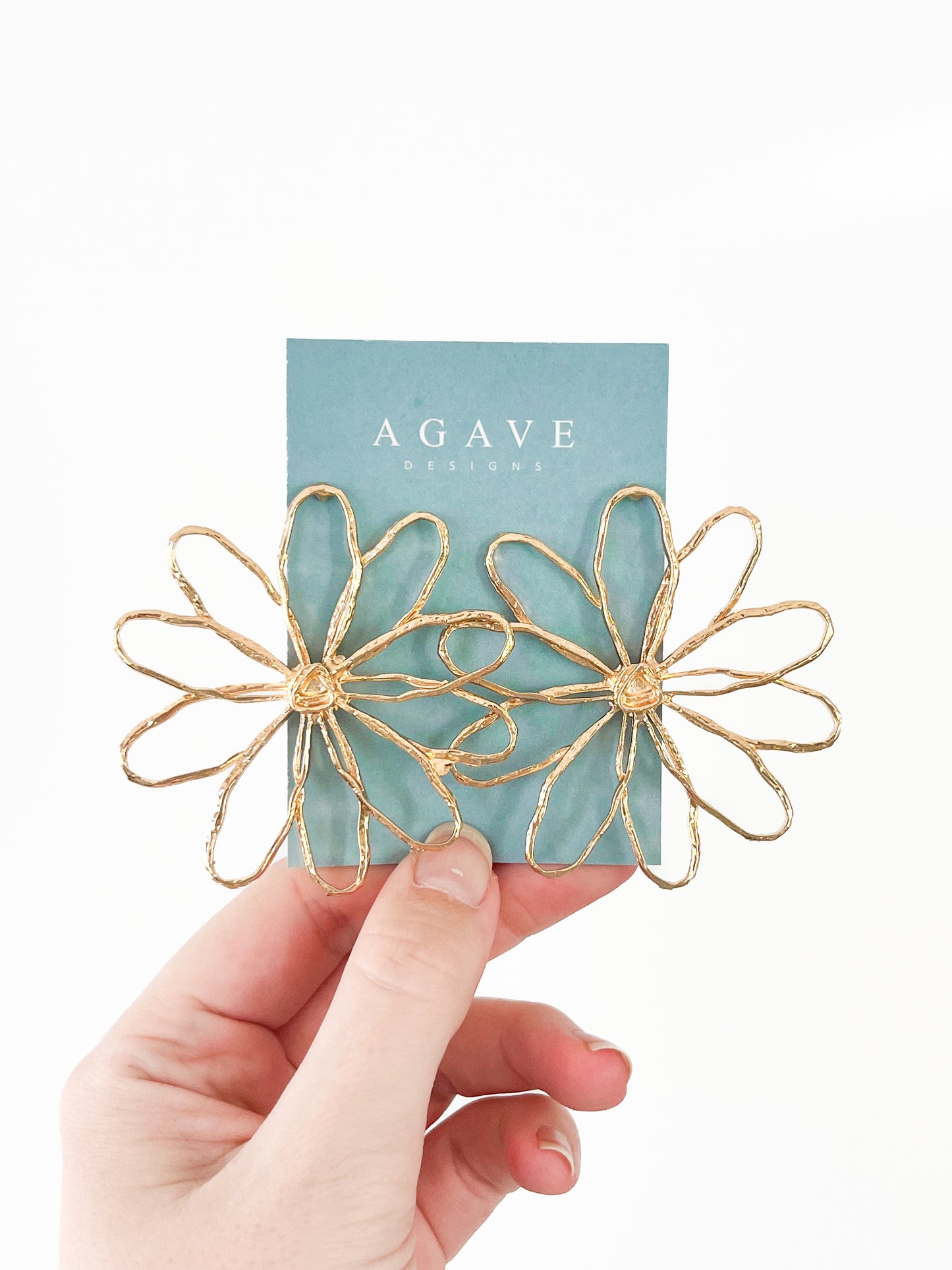 A hand holds a blue card with "Agave Designs" printed on it, displaying the Large Daisy Studs—two flower-shaped earrings crafted from zinc alloy featuring delicate petal details. The plain white background beautifully highlights the elegant design and color contrast.