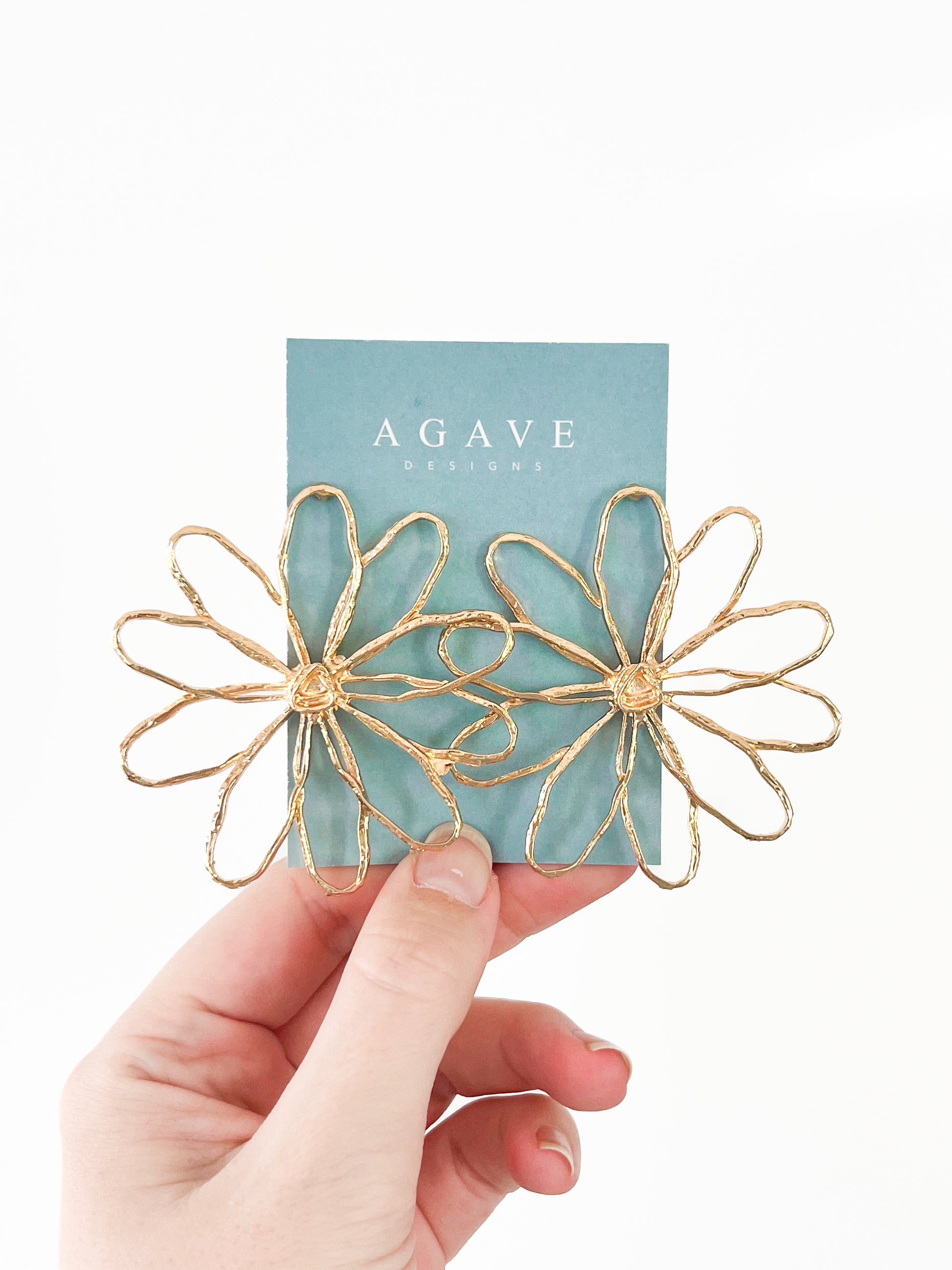 A hand holds a blue card with "Agave Designs" printed on it, displaying the Large Daisy Studs—two flower-shaped earrings crafted from zinc alloy featuring delicate petal details. The plain white background beautifully highlights the elegant design and color contrast.