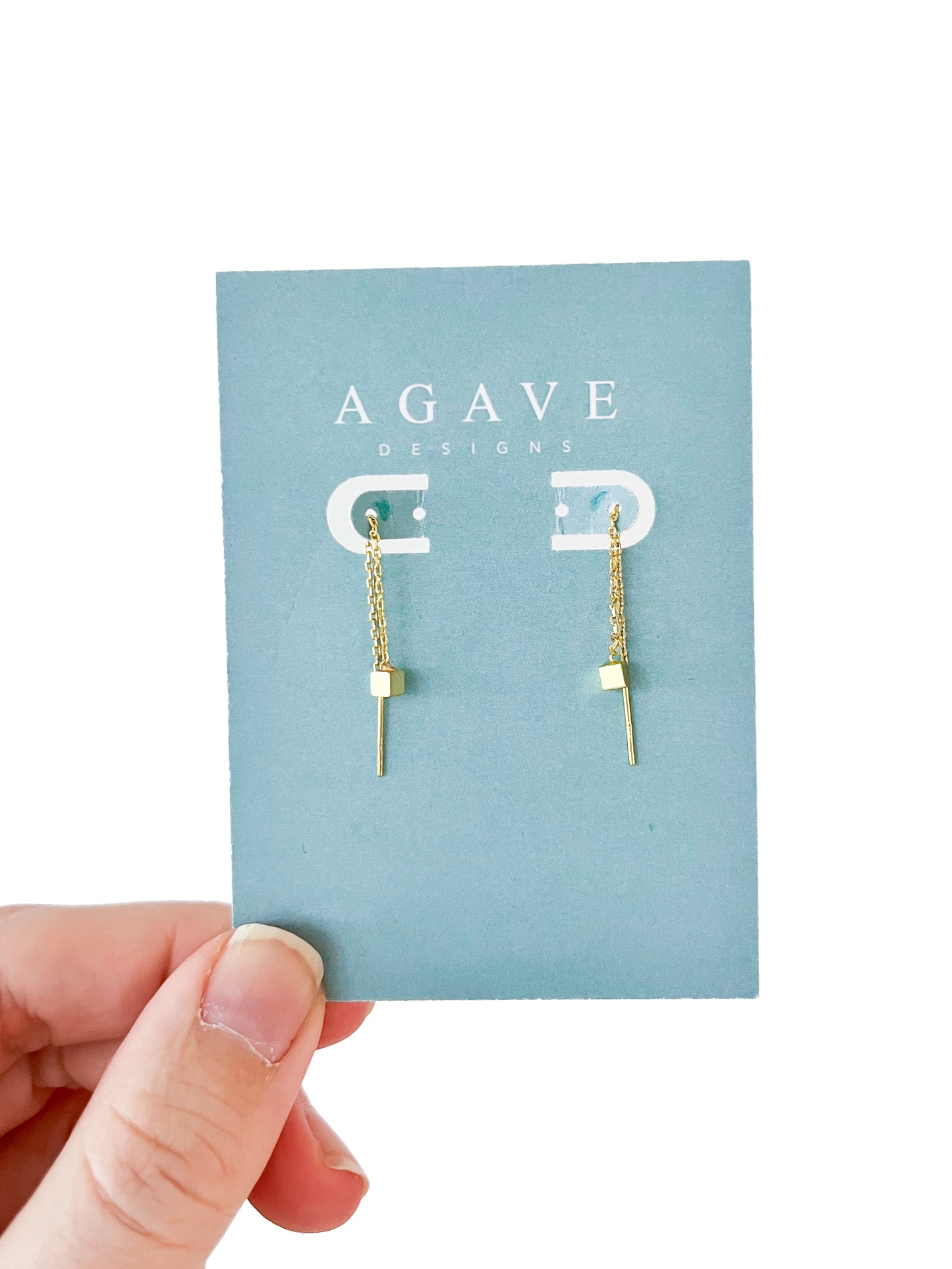 A hand presents a card from "Agave Designs" displaying the "Little Square danglie Gold" earrings, crafted in gold color zinc alloy with a chain featuring a cube and thin bar charm, all on a plain white background.