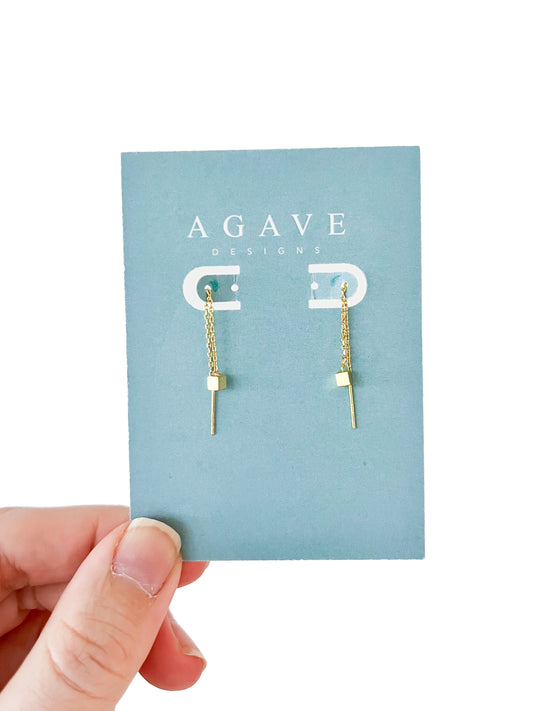 A hand presents a card from "Agave Designs" displaying the "Little Square danglie Gold" earrings, crafted in gold color zinc alloy with a chain featuring a cube and thin bar charm, all on a plain white background.