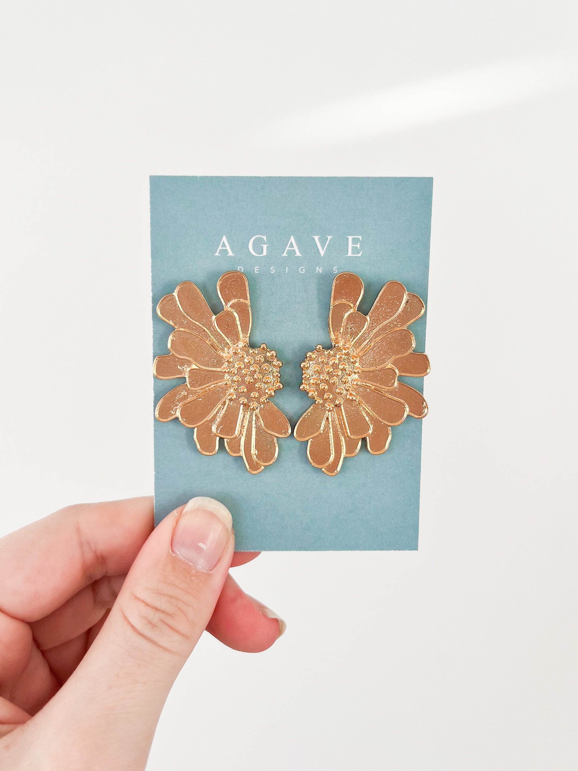 A hand presents a pair of gold floral earrings crafted from zinc alloy against a teal card labeled "Agave Designs." These "Big Flower Studs" epitomize elegance and craftsmanship.