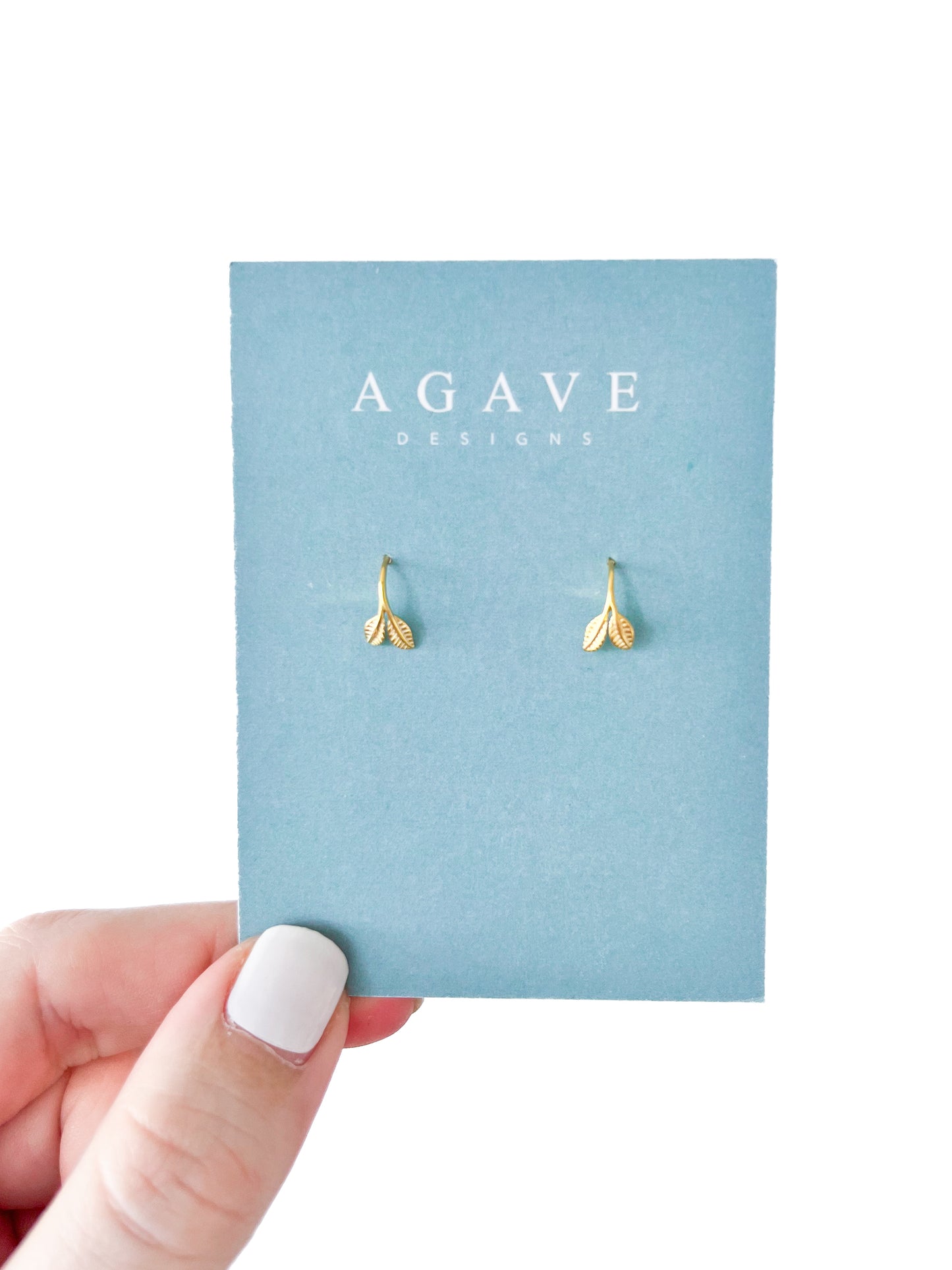 A hand with crisp white nail polish holds a blue card reading "AGAVE DESIGNS," displaying the Baby Leaf Huggies, elegant leaf-shaped gold earrings.