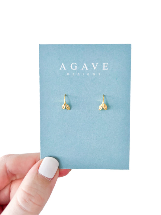 A hand with crisp white nail polish holds a blue card reading "AGAVE DESIGNS," displaying the Baby Leaf Huggies, elegant leaf-shaped gold earrings.