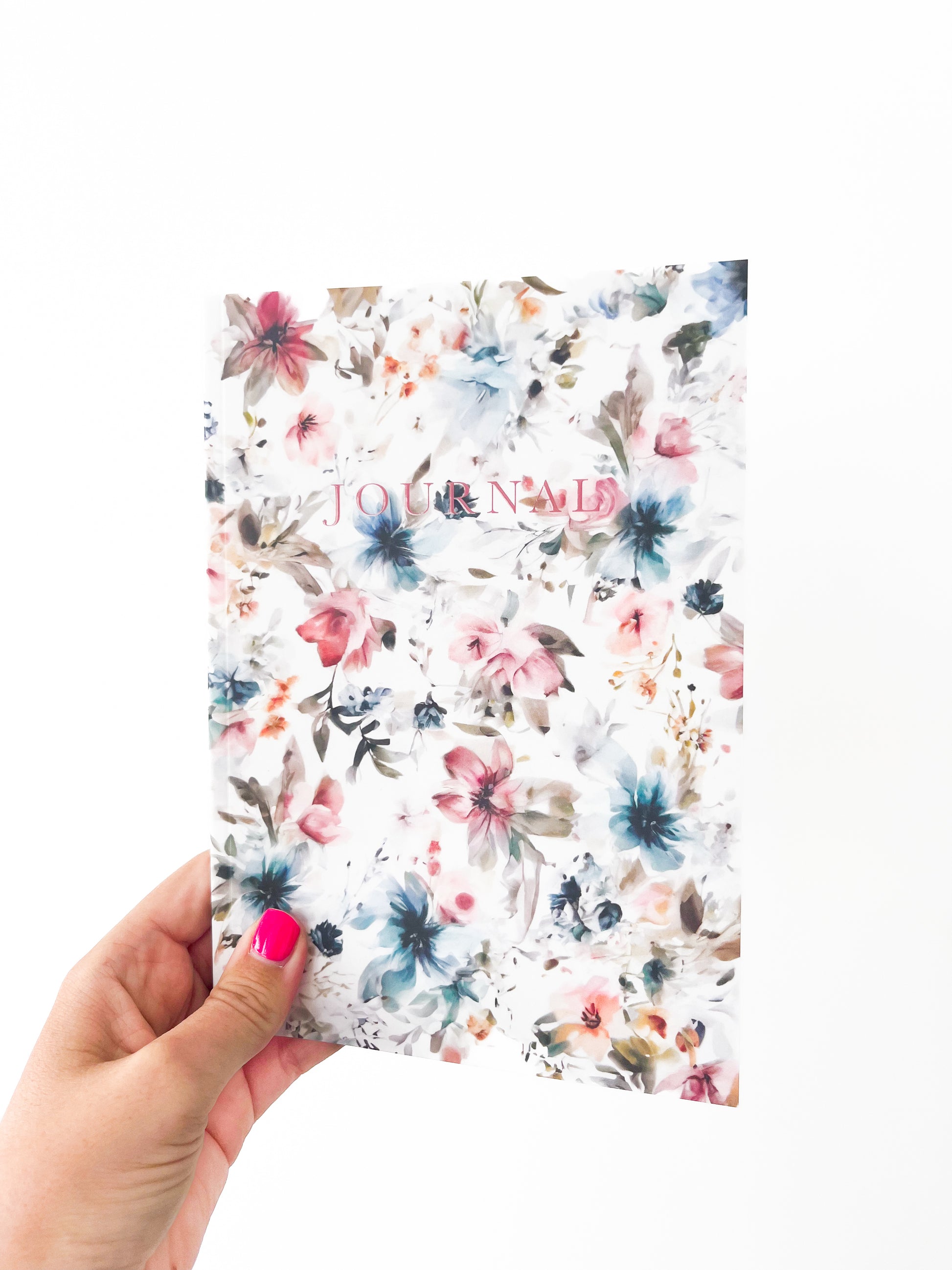 A hand with pink nail polish holds a floral-patterned notebook named "March Notebook" by Agave Designs, with the word "JOURNAL" written on the cover in pink lettering. This charming paperback journal features thick paper and lined pages, perfect for all your notes. The background of the image is white.