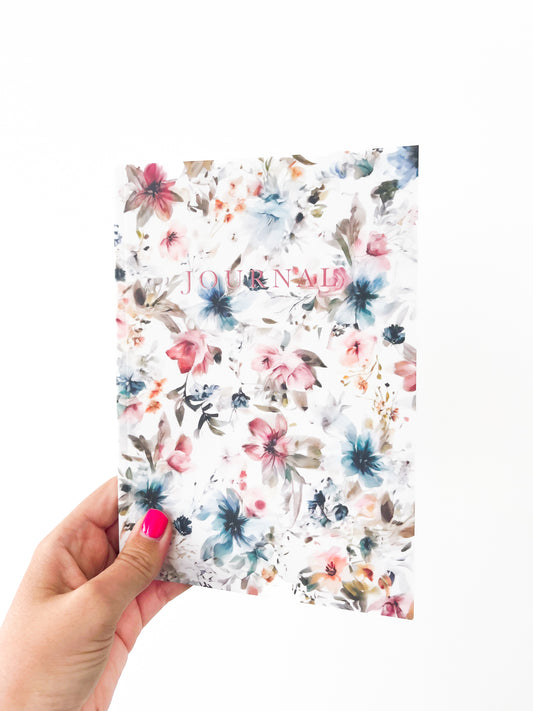 A hand with pink nail polish holds a floral-patterned notebook named "March Notebook" by Agave Designs, with the word "JOURNAL" written on the cover in pink lettering. This charming paperback journal features thick paper and lined pages, perfect for all your notes. The background of the image is white.