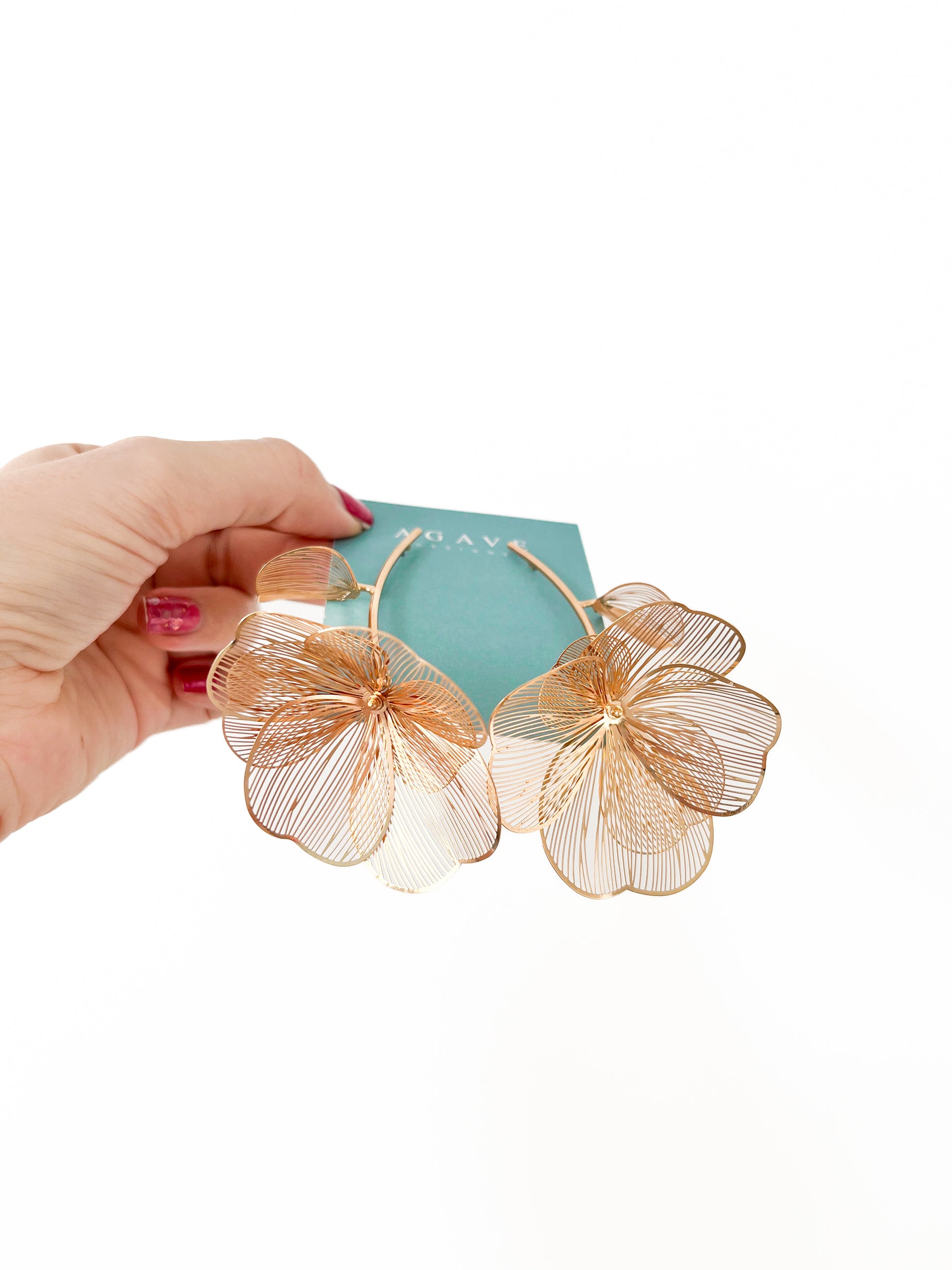A hand with red nail polish elegantly holds a pair of Chunky Gold Flower Danglies, crafted by Agave Designs from zinc alloy and measuring 85mm x 60mm, against a white background.