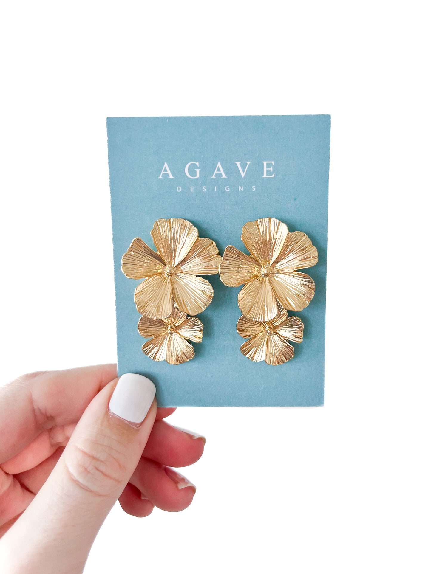 A hand with white-painted nails holds a blue card showing Agave Designs' Double Flower Studs, crafted in stainless steel. These gold-colored floral earrings feature three layered petals, measuring 40mm x 28mm for a textured, elegant look.