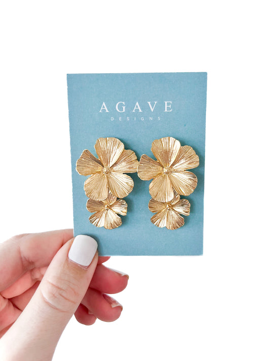 A hand with white-painted nails holds a blue card showing Agave Designs' Double Flower Studs, crafted in stainless steel. These gold-colored floral earrings feature three layered petals, measuring 40mm x 28mm for a textured, elegant look.