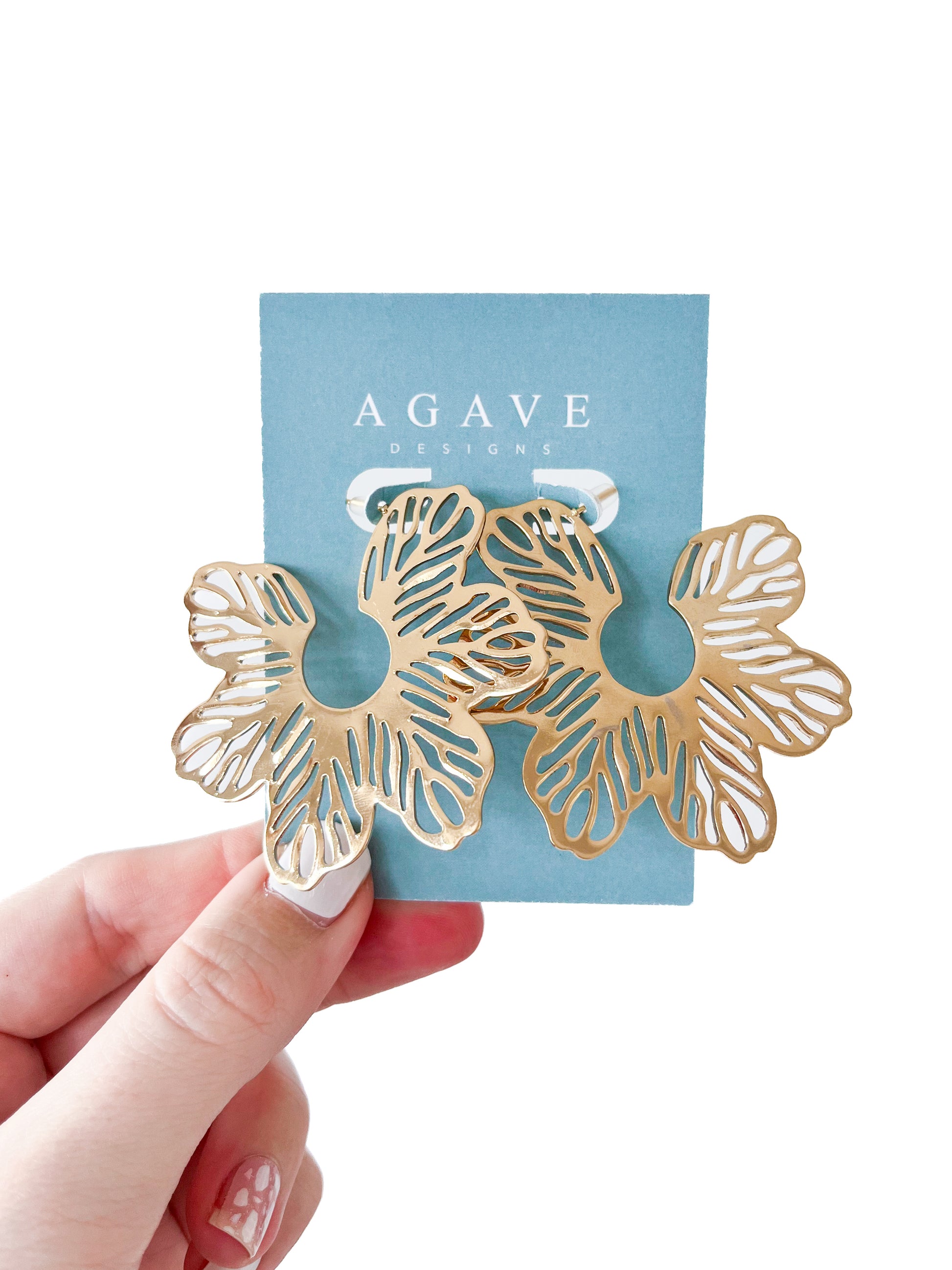 Hand holds gold Flower Studs earring on a light blue card labeled "Agave Designs." The 60mm stainless steel earrings feature cut-out details creating an intricate floral pattern.