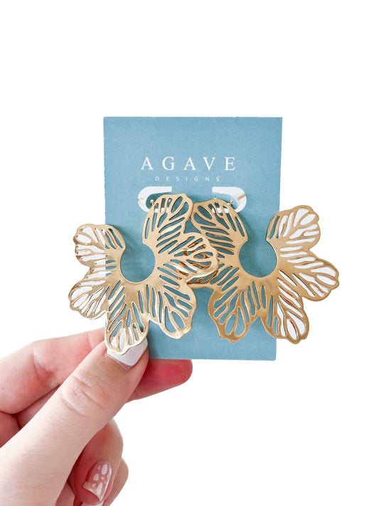 Hand holds gold Flower Studs earring on a light blue card labeled "Agave Designs." The 60mm stainless steel earrings feature cut-out details creating an intricate floral pattern.