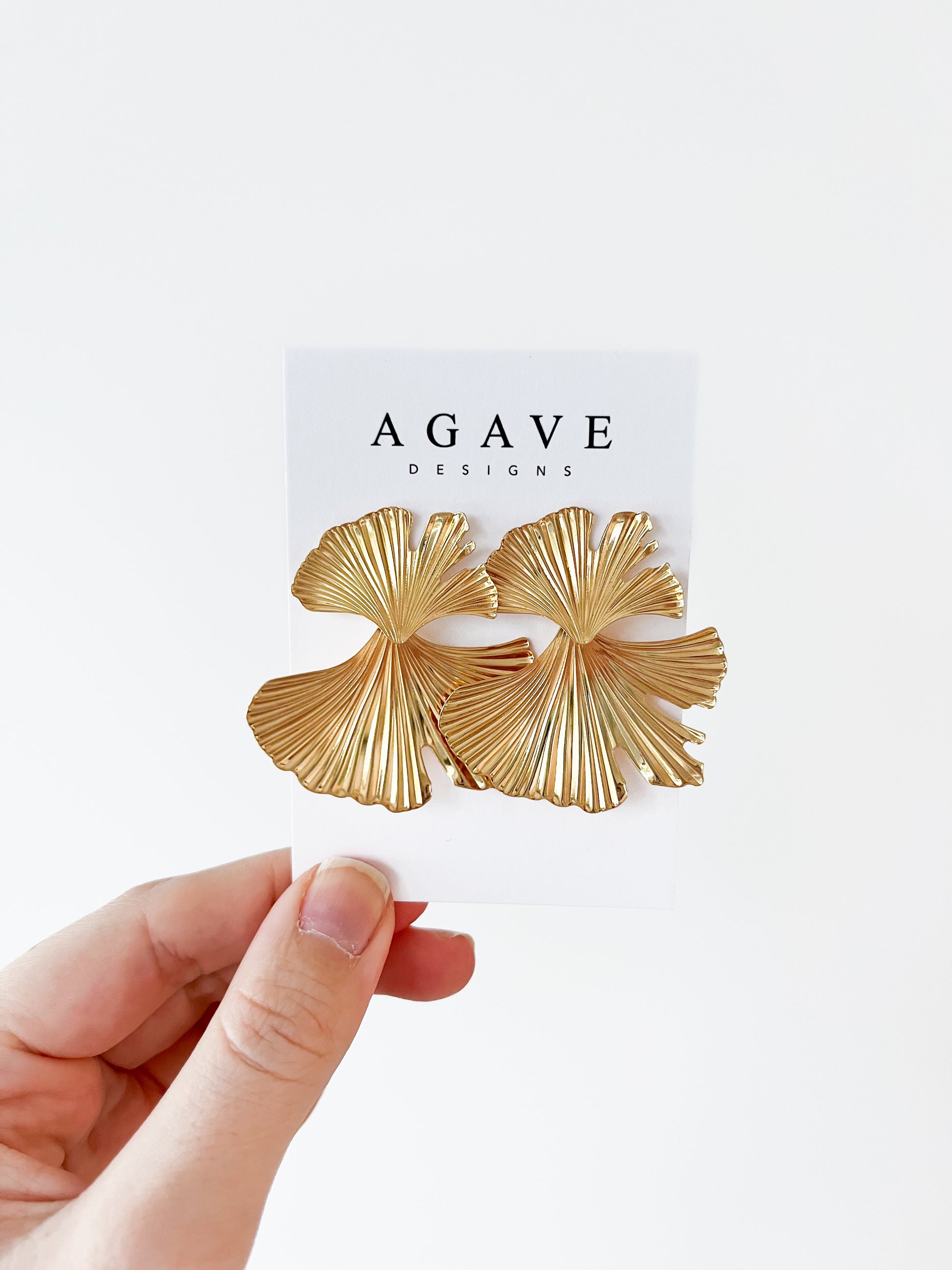 A hand displays a Ginkgo Danglies card featuring large, gold, fan-shaped earrings with a leaf-like zinc alloy design. "Agave Designs" is printed at the top against a plain white wall background.