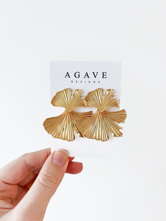 A hand displays a Ginkgo Danglies card featuring large, gold, fan-shaped earrings with a leaf-like zinc alloy design. "Agave Designs" is printed at the top against a plain white wall background.