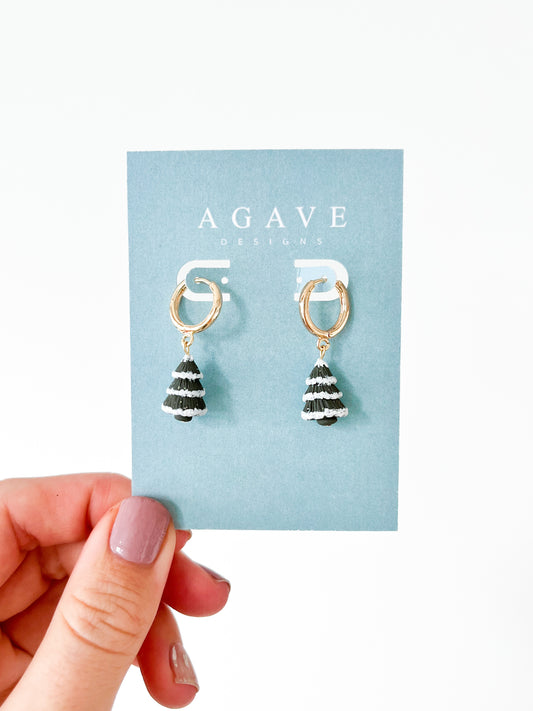 A hand with painted nails holds a blue Agave Designs card showcasing the Christmas Tree Huggies. These gold plated hoop earrings feature a dangling black and white striped cone-shaped charm, striking the perfect balance between size and style.