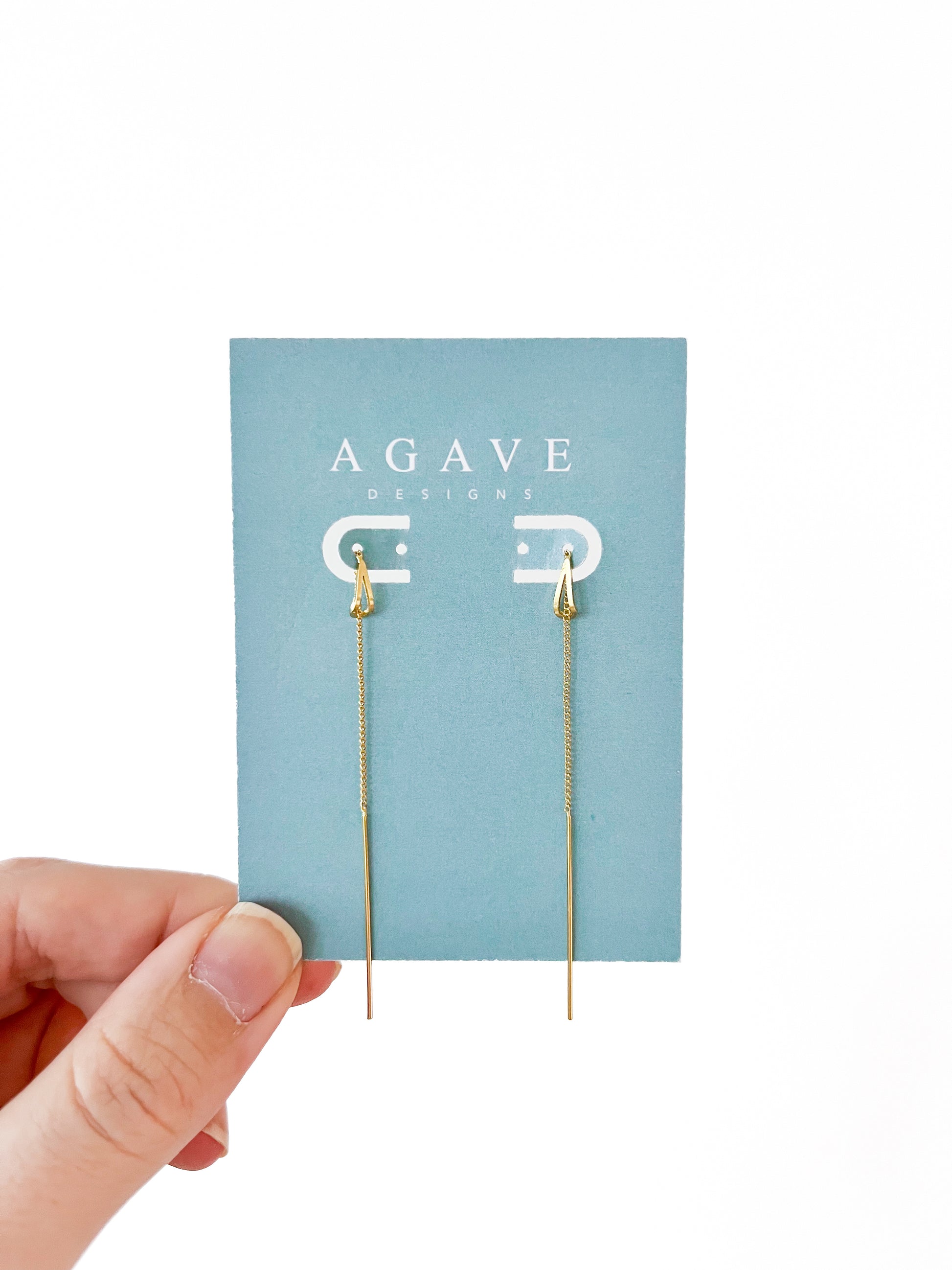 A hand holds a blue "Agave Designs" card displaying Drop Ear line threader danglies. These exquisite gold earrings feature delicate chains and arrowhead shapes, creating a sleek design that stands out against the white background, highlighting their elegance and charm.