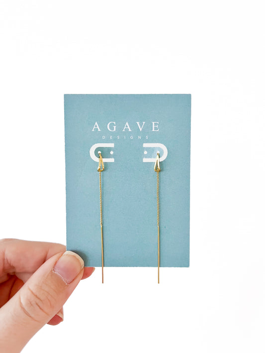 A hand holds a blue "Agave Designs" card displaying Drop Ear line threader danglies. These exquisite gold earrings feature delicate chains and arrowhead shapes, creating a sleek design that stands out against the white background, highlighting their elegance and charm.
