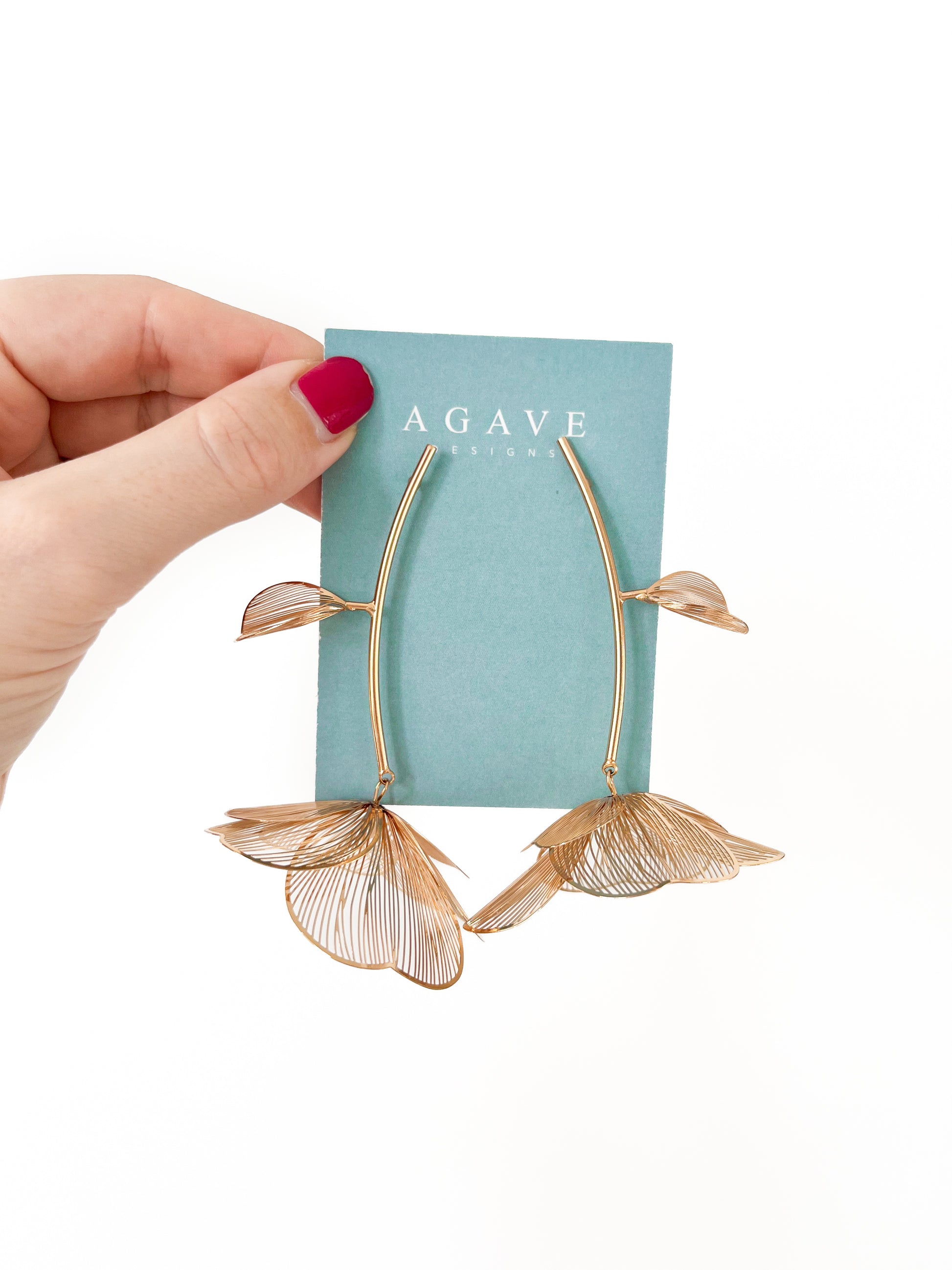 A hand with pink nail polish holds a pair of "Chunky Gold Flower Danglies" against a teal card labeled "Agave Designs." These delicate earrings, measuring 85mm x 60mm, feature large petal-like designs with a subtle wireframe structure.