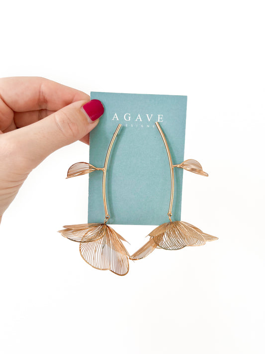 A hand with pink nail polish holds a pair of "Chunky Gold Flower Danglies" against a teal card labeled "Agave Designs." These delicate earrings, measuring 85mm x 60mm, feature large petal-like designs with a subtle wireframe structure.