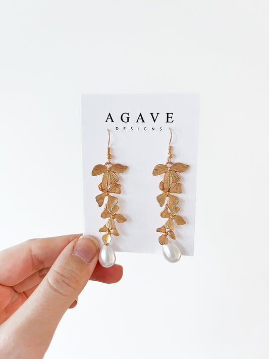 A hand showcases the exquisite "Long Floral Pearl Danglies" by Agave Designs against a white backdrop, crafted from gold zinc alloy in a graceful leaf design with a pearl-like bead, elegantly presented on an "AGAVE DESIGNS" card.