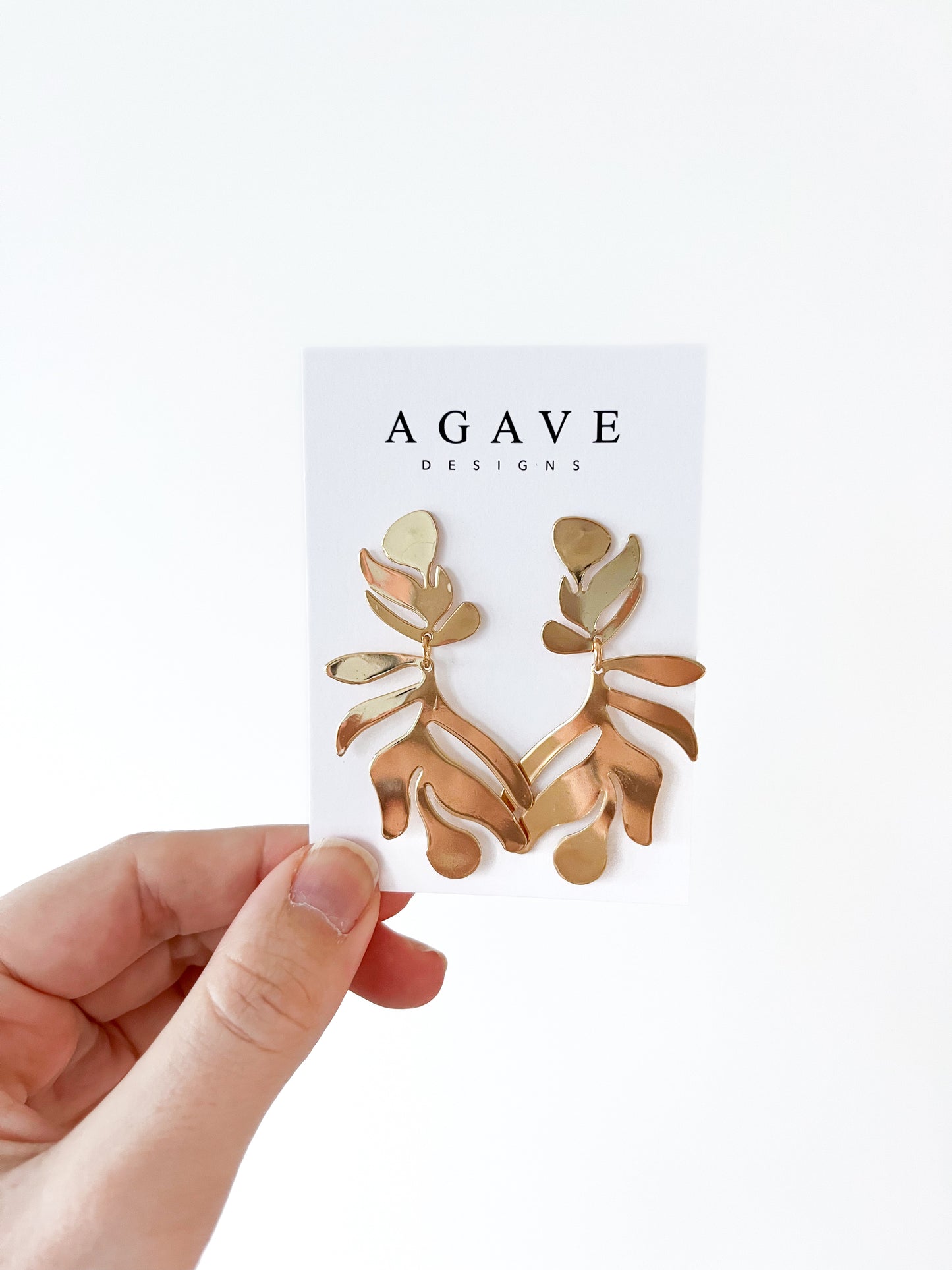 A hand displays a card with the "Agave Designs" brand, presenting exquisite gold-toned geometric leaf danglies made from high-quality zinc alloy against a pristine white background.