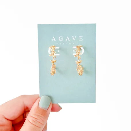 A hand with green-painted nails holds a blue card displaying a pair of 14k gold plated "Floral Gold Hoops" from Agave Designs. The 27mm earrings feature delicate flower and leaf details arranged in a vertical line.