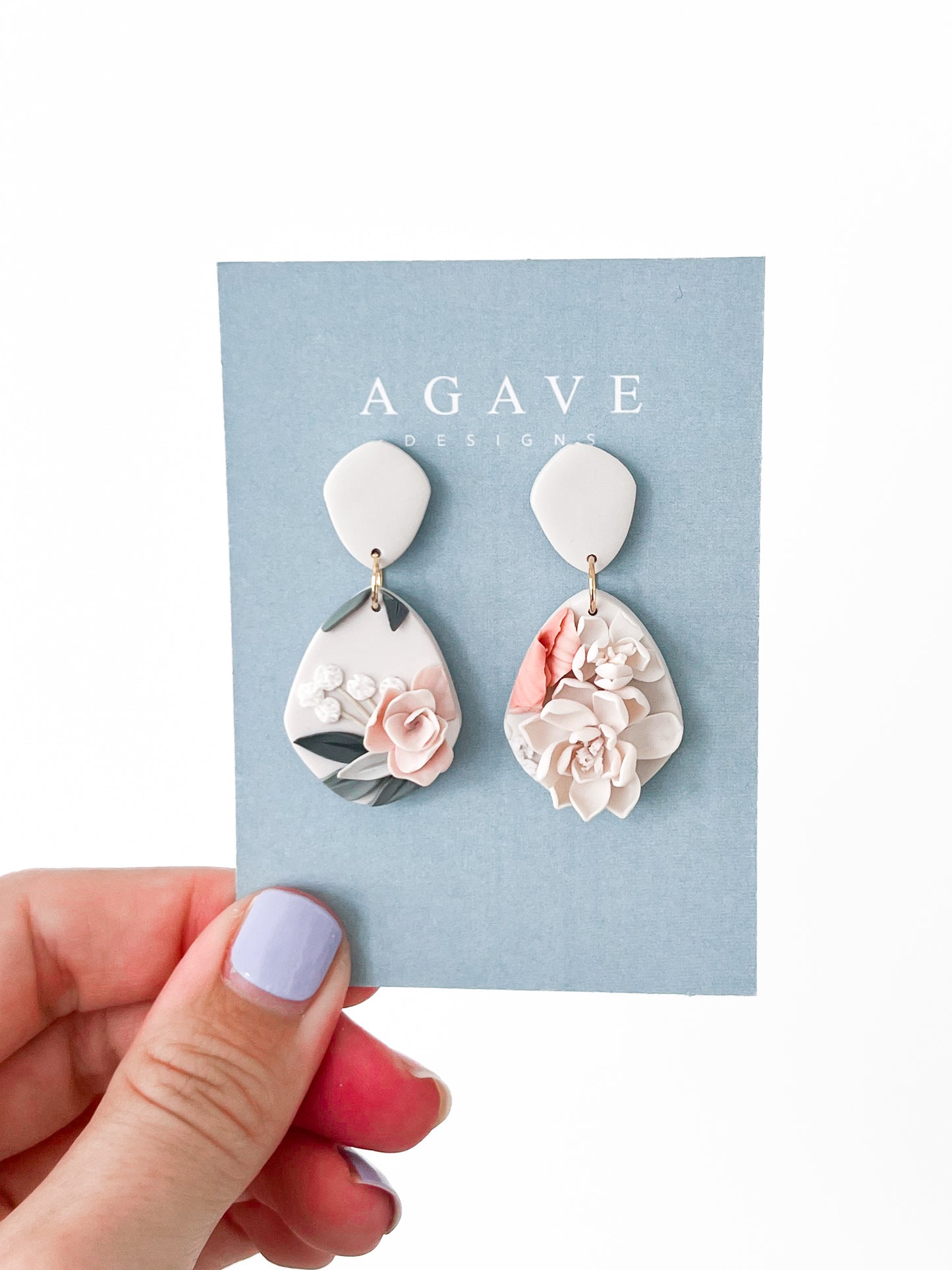 A hand with lavender nail polish holds a blue "Agave Designs" card showing the Tammy earrings: 40mm x 22mm sterling silver teardrop earrings with raised floral designs in soft pink, white, gray, and sage.