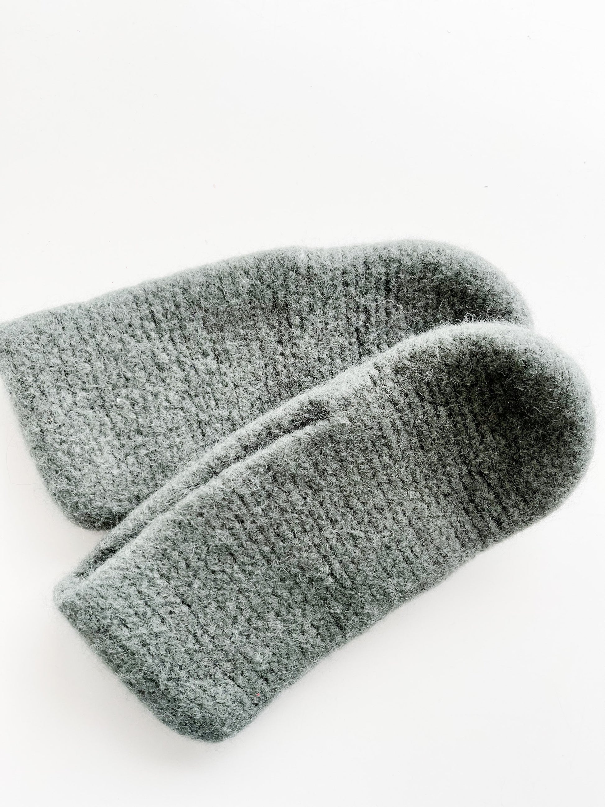 A close-up image of a pair of sage green wool slippers made from luxurious Norwegian wool, set against a white background. The slippers, crafted with Agave Designs yarn, showcase a soft and fuzzy texture with a knitted pattern visible across their surface, positioned slightly overlapping each other.