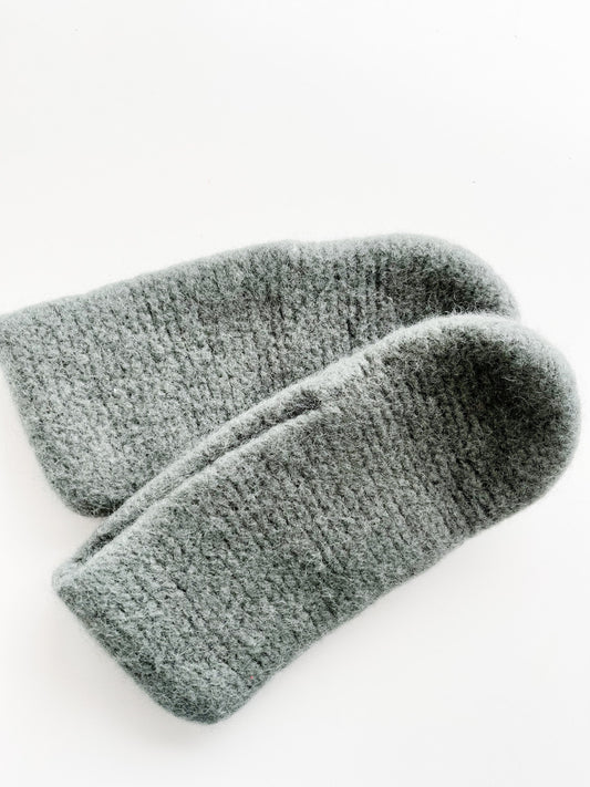 A close-up image of a pair of sage green wool slippers made from luxurious Norwegian wool, set against a white background. The slippers, crafted with Agave Designs yarn, showcase a soft and fuzzy texture with a knitted pattern visible across their surface, positioned slightly overlapping each other.