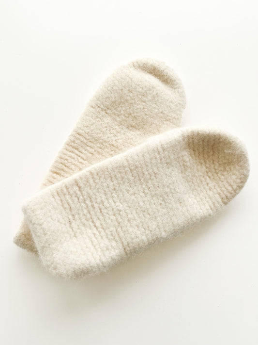 On a white background lies a pair of Cream Wool Slippers from Agave Designs. These cream-colored, woolen slipper socks are crafted from Norwegian wool and boast a soft, fuzzy texture with thick fabric, promising warmth and comfort. For optimal care, it is recommended to hand wash them gently.