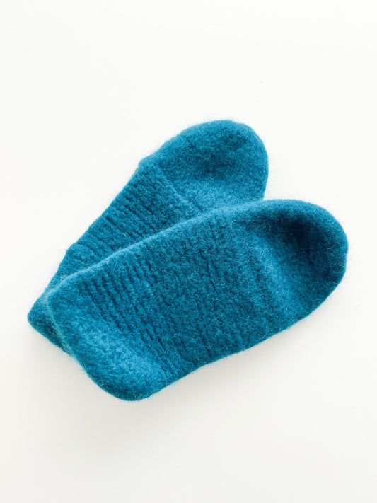 A pair of teal/turquoise slippers, made from luxurious Norwegian wool by Agave Designs, is neatly positioned on a plain white background. The slippers have a fuzzy texture and appear soft and cozy, ideal for providing warmth. Remember to hand wash to preserve their quality.