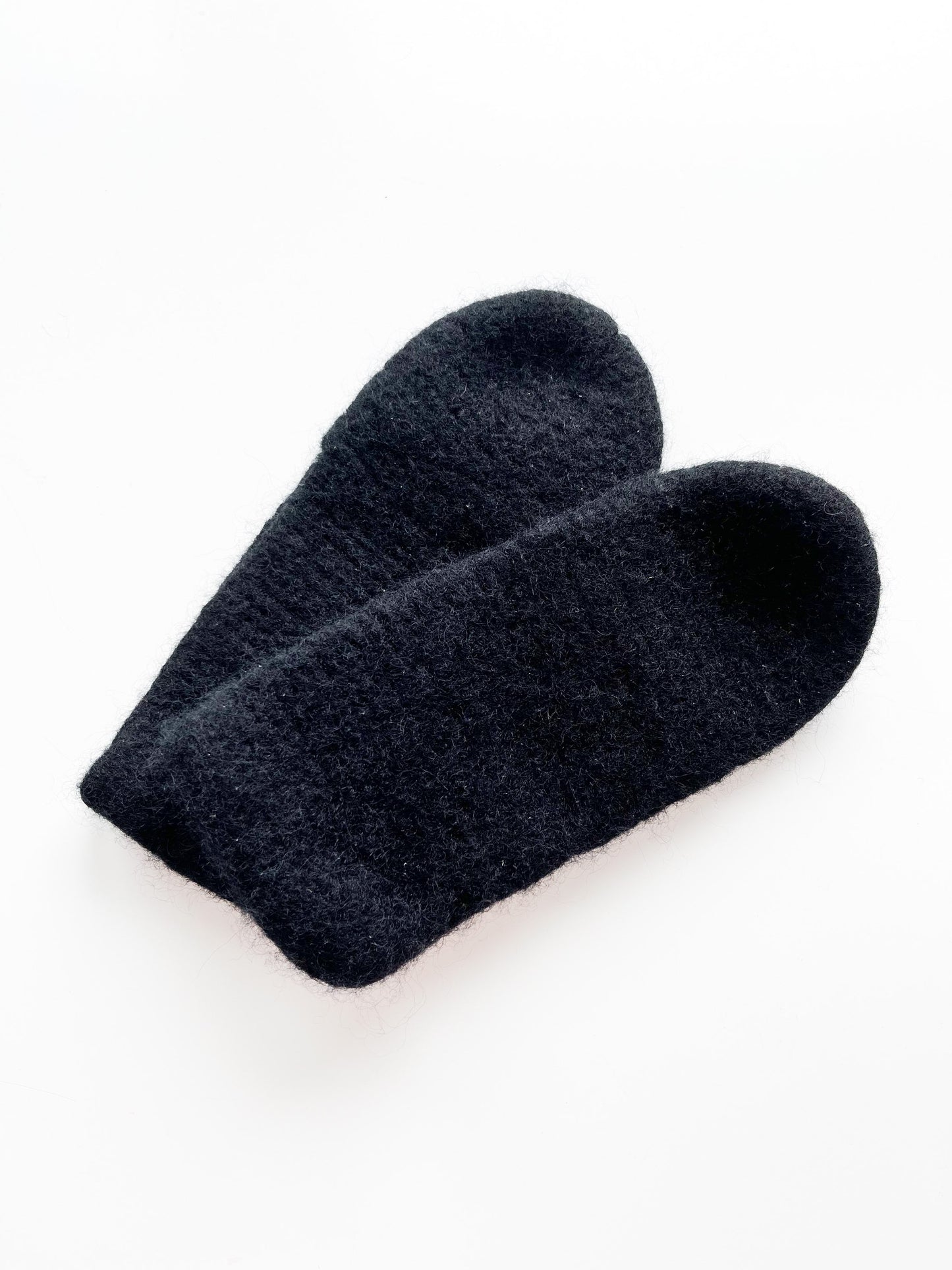 A pair of black, woolen slippers made by Agave Designs is laid flat on a white surface. The slippers appear soft and are designed without any visible patterns or decorations, with a tag advising to hand wash separately.