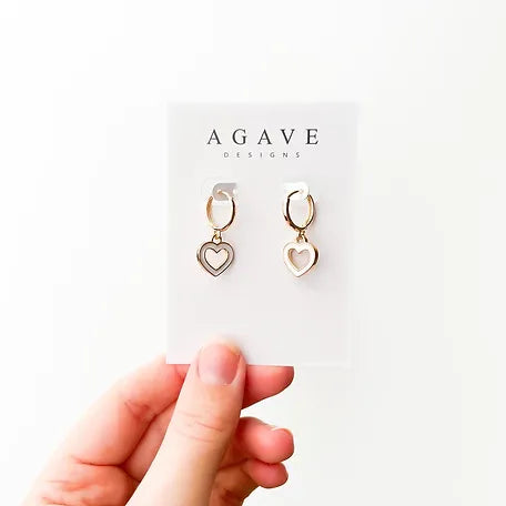 A hand is shown holding a white card from "Agave Designs," displaying a pair of Heart Huggies. These gold-plated, heart-shaped earrings measure 25mm x 14mm and feature small hoops with dangling hollow hearts. The background is plain white.