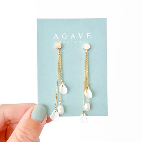 A hand with mint green nail polish holding a blue card labeled "Agave Designs." The card displays the Josefine 14k gold plated dangle earrings, featuring thin chains and white teardrop-shaped beads.