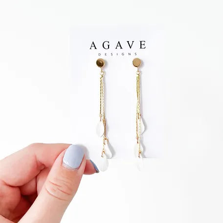 A hand with light blue nail polish holds a pair of elegant handmade 3 petal drop earrings, named "Nora," against a white background. The earrings, by Agave Designs, feature gold plated chains and white teardrop-shaped pendants hanging from circular gold studs.