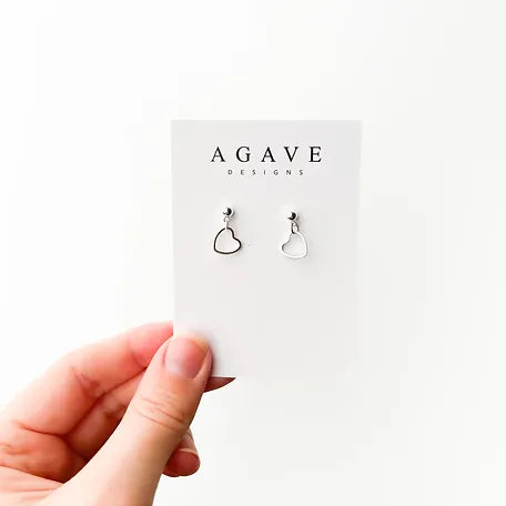 A hand holds a card displaying a pair of delicate Simple Heart Danglies, sterling silver earrings. The card is labeled "Agave Designs" at the top. The background is plain white.