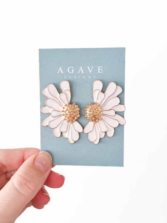 A hand holds a sleek card displaying "Agave Designs," showcasing two exquisite White ‘n Gold Flower Studs with radiant gold centers. The jewelry shines vividly against the plain white background.