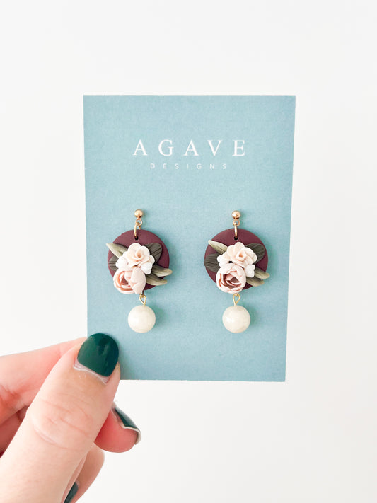 A hand with forest green-painted nails holds a card displaying the Daphne earrings from Agave Designs. The gold-plated earrings feature a circular design with floral elements and a dangling pearl-like bead. The card has a turquoise background and the brand's name in white text at the top.